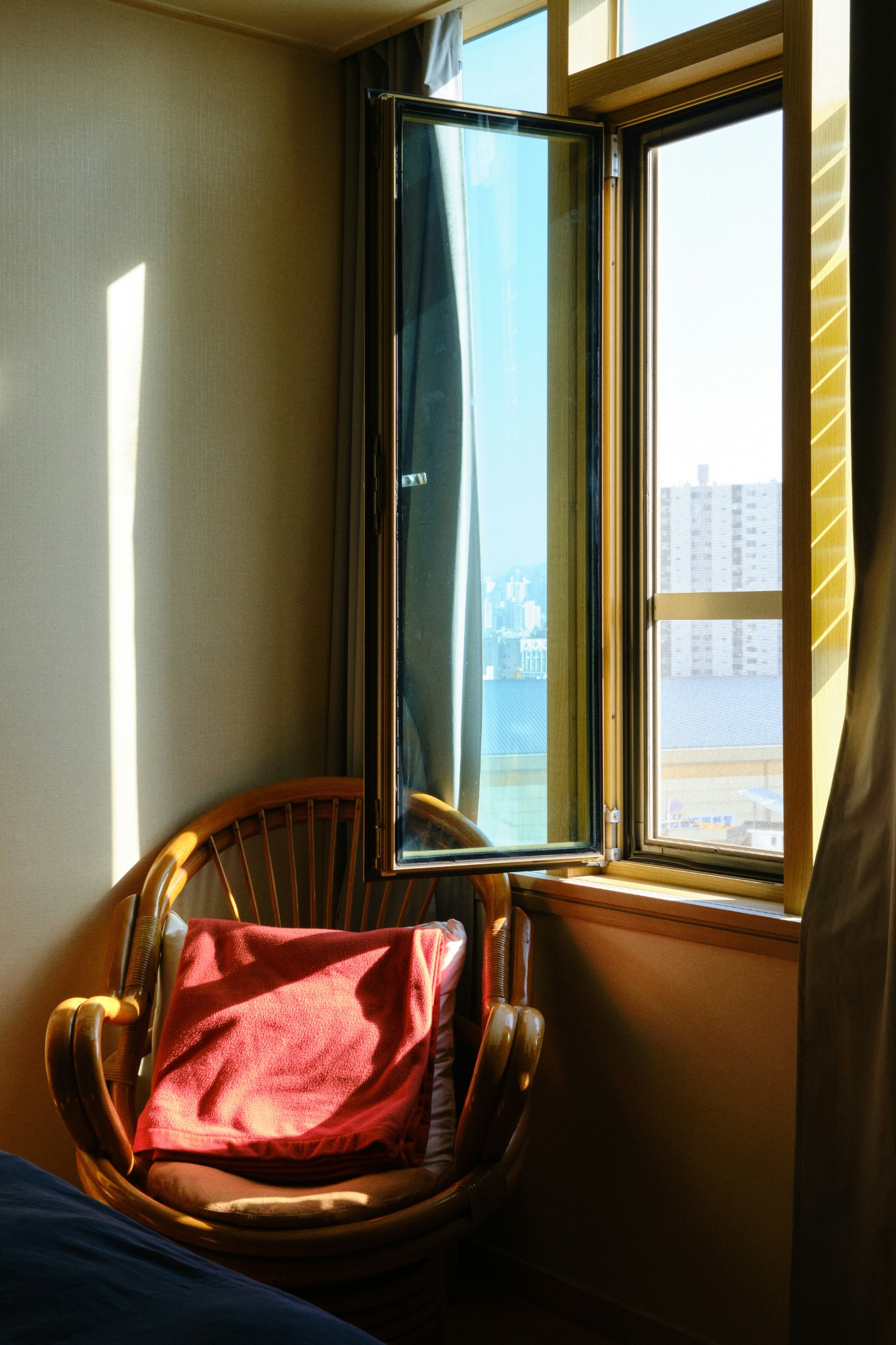 Poltrona de rattan com almofada vermelha junto a uma janela aberta, iluminada pelo sol.